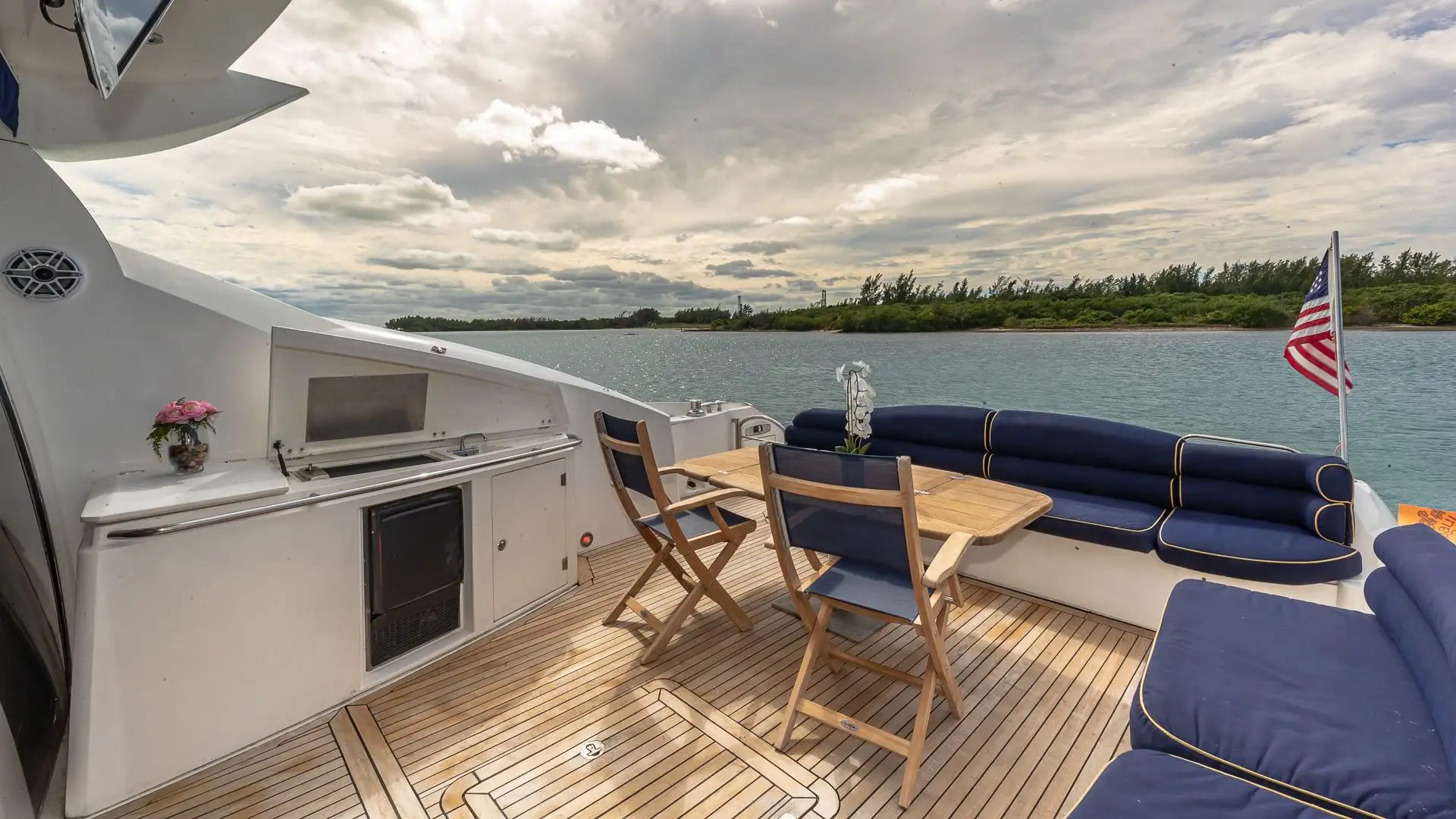 Boat deck with seating, wooden table, grill, and ocean view under cloudy sky.