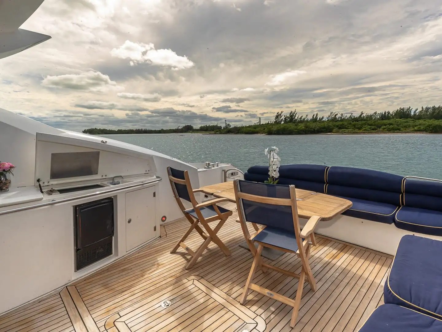 Boat deck with seating, wooden table, grill, and ocean view under cloudy sky.