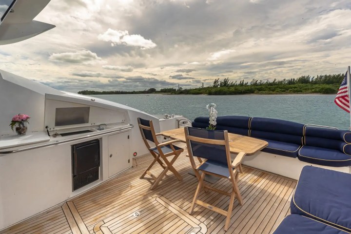Boat deck with seating, wooden table, grill, and ocean view under cloudy sky.