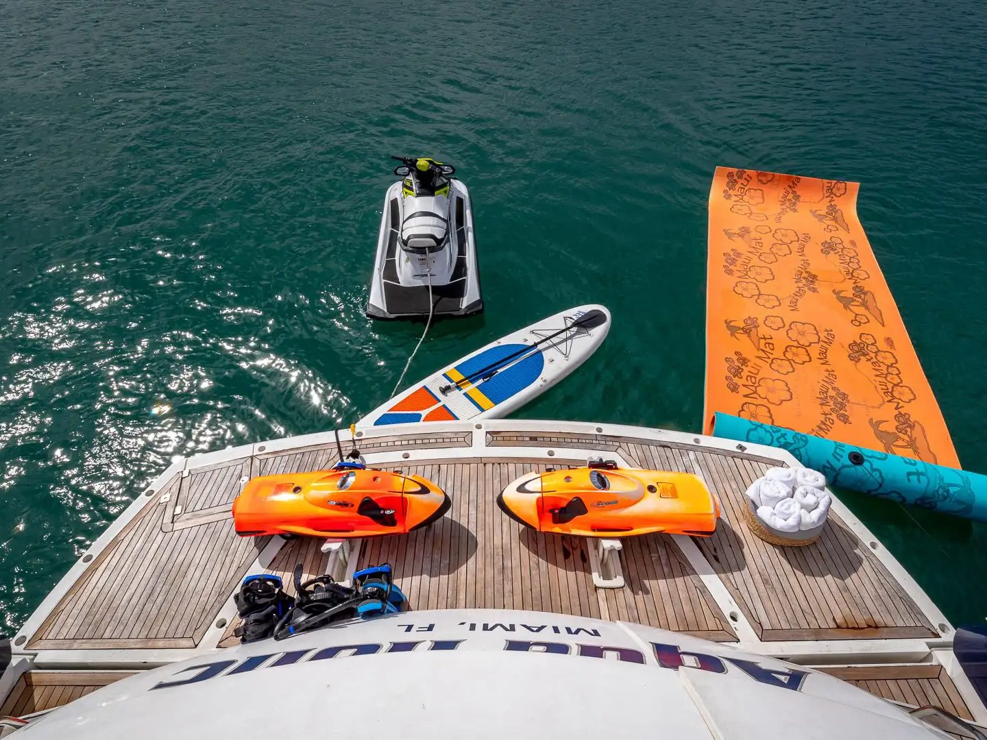 View from yacht deck with jet ski, paddleboard, and orange mat floating on water.