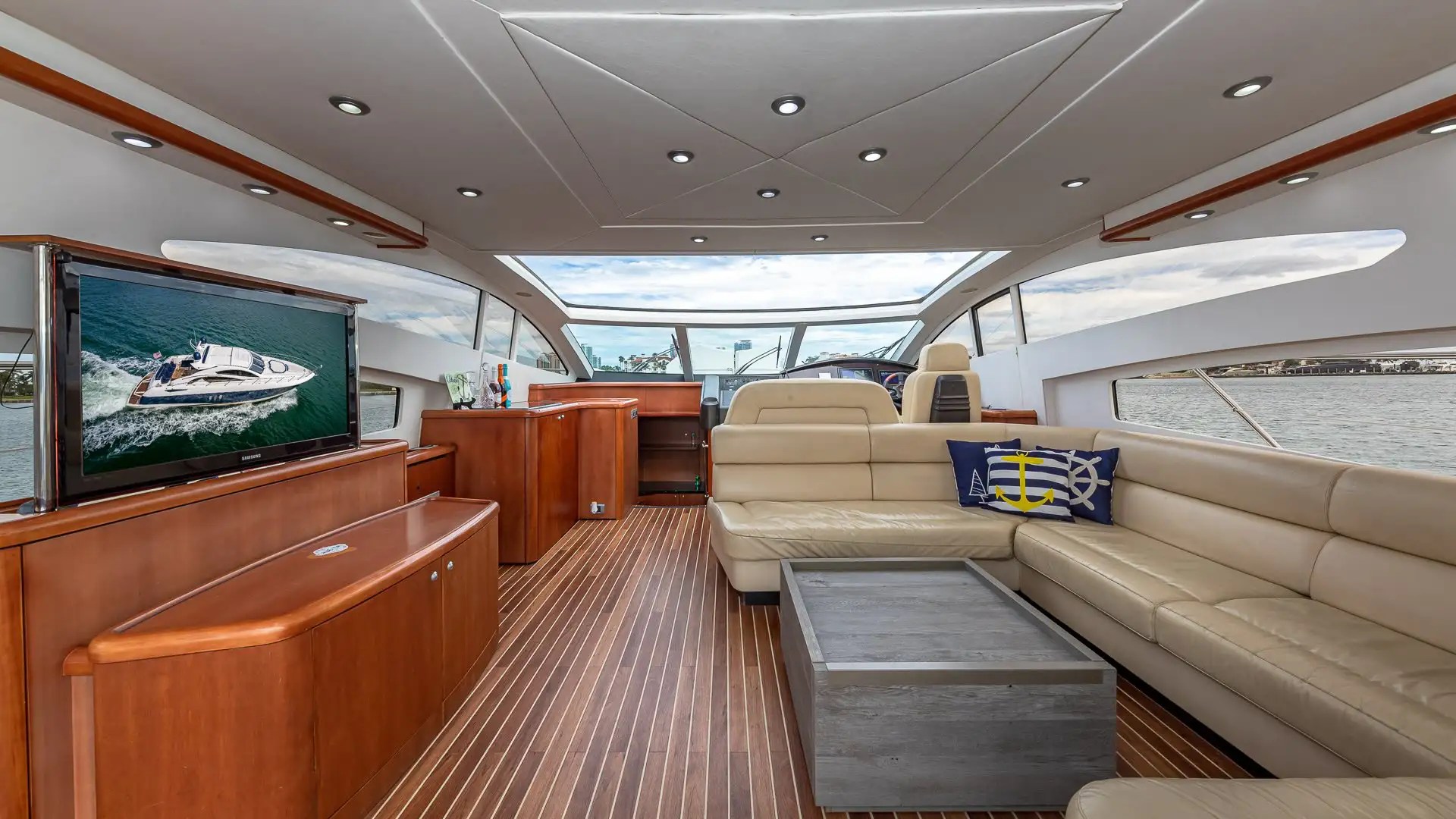Luxury yacht interior with leather seating, wooden flooring, TV, and large windows showing a view of the water.