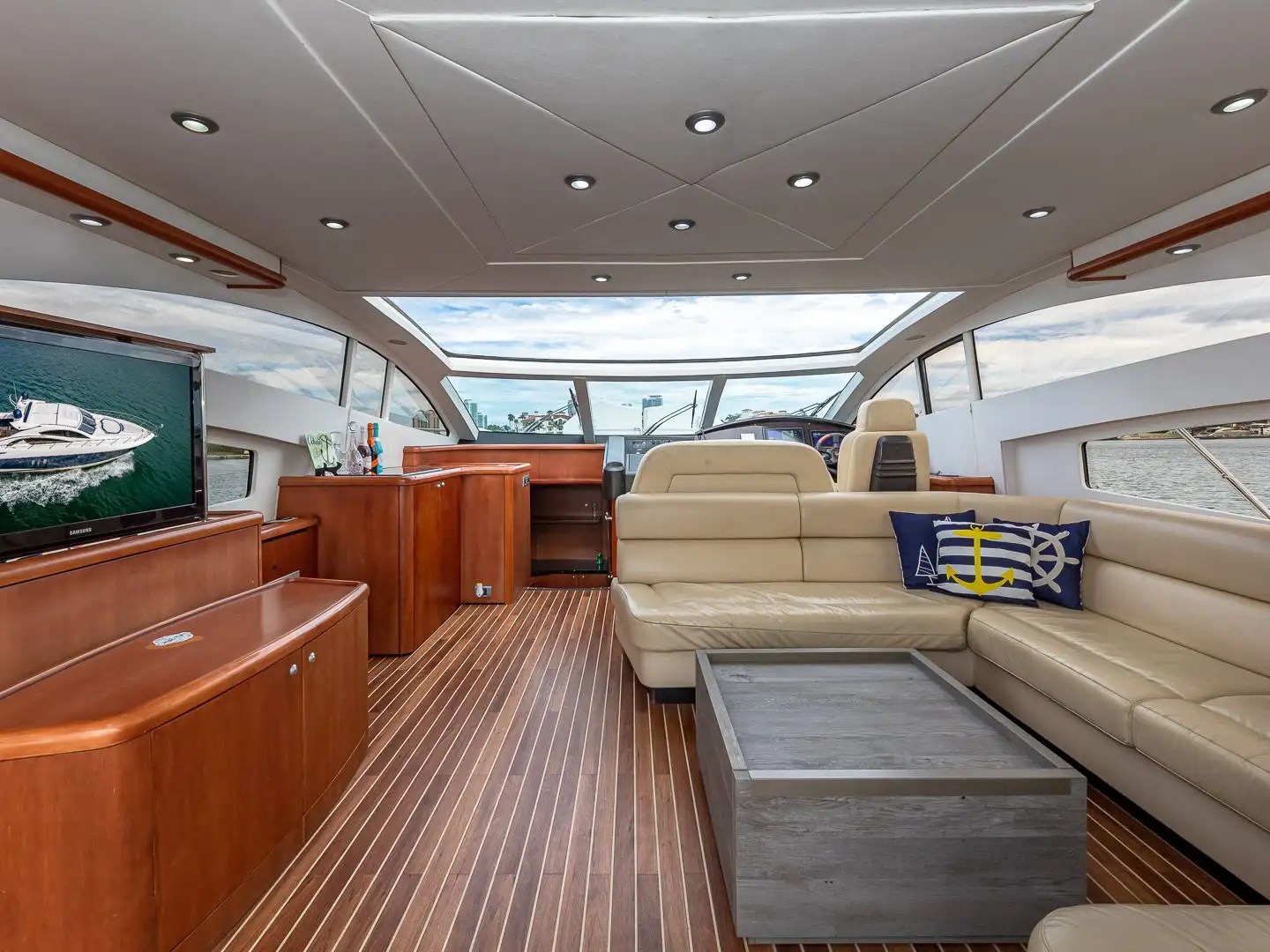 Luxury yacht interior with leather seating, wooden flooring, TV, and large windows showing a view of the water.