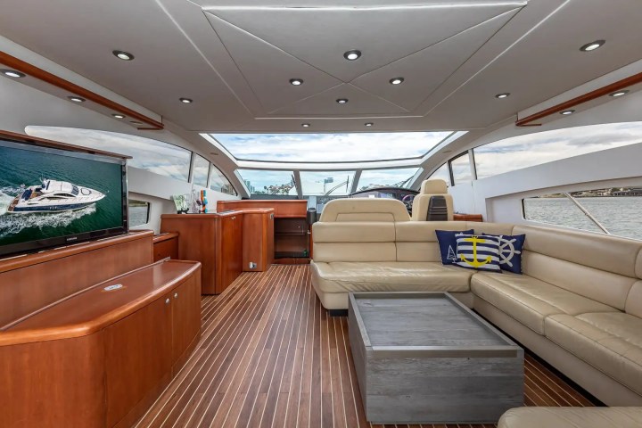Luxury yacht interior with leather seating, wooden flooring, TV, and large windows showing a view of the water.