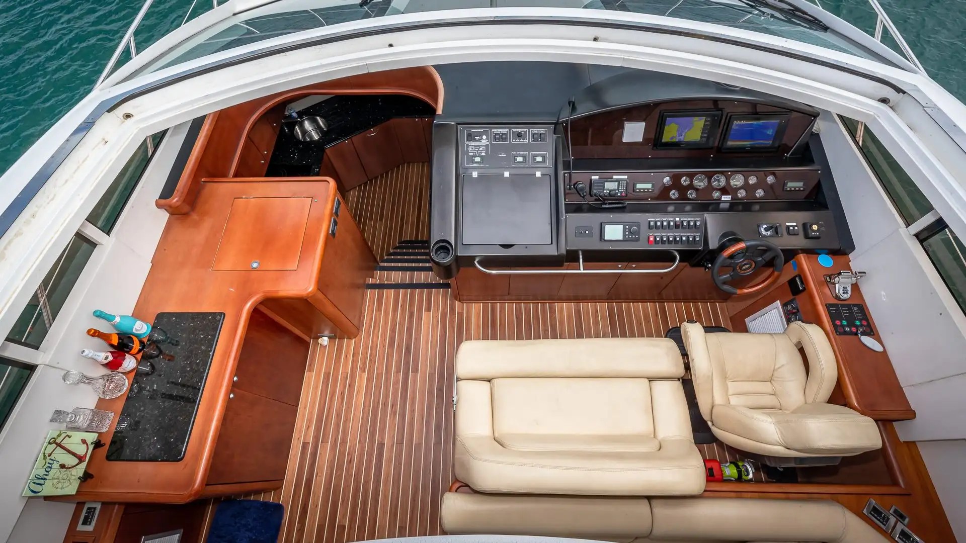 Boat cockpit with steering wheel, control panels, and beige seating on polished wood floor.