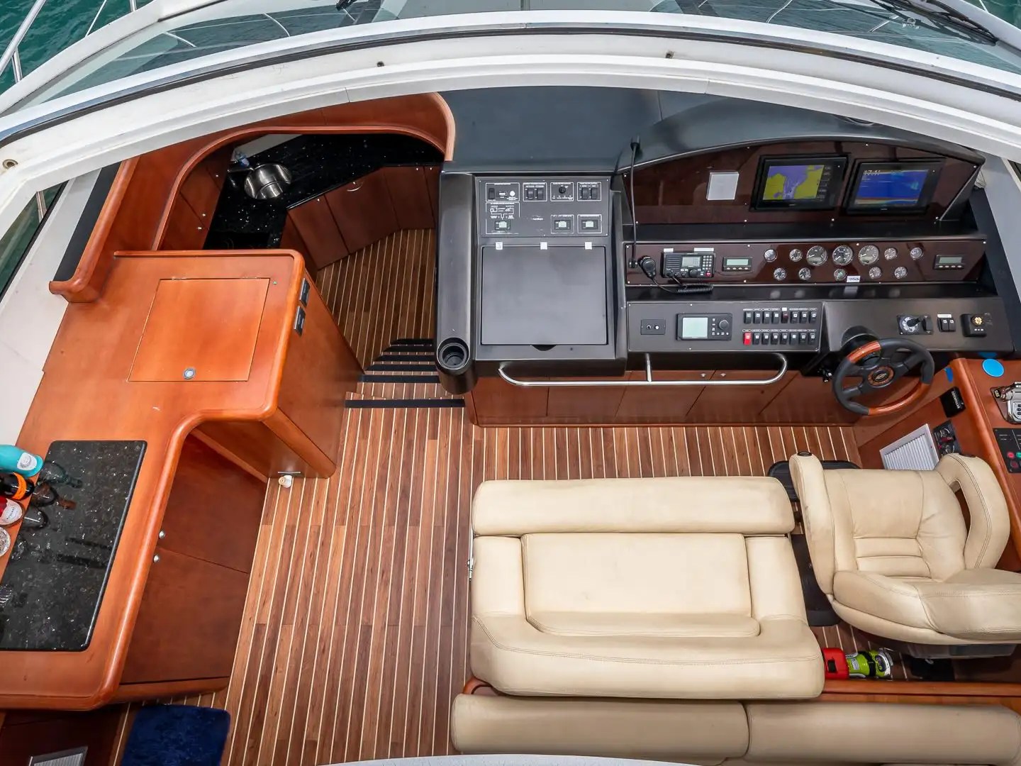 Boat cockpit with steering wheel, control panels, and beige seating on polished wood floor.