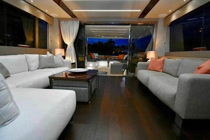 Luxurious yacht interior with gray sofas, orange cushions, and dark wood floor, lit by lamps.