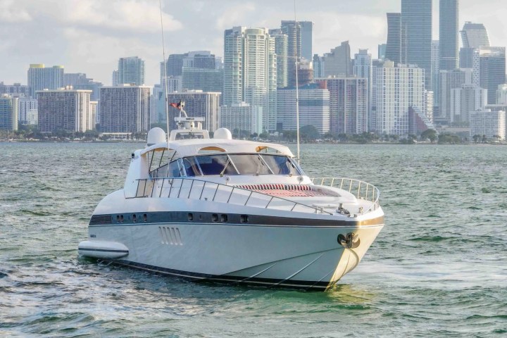White yacht sailing with a city skyline in the background under a partly cloudy sky.