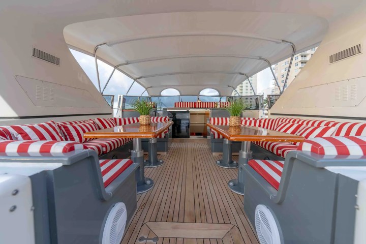 Open yacht deck with red-striped seating and wooden tables, covered by a canopy.