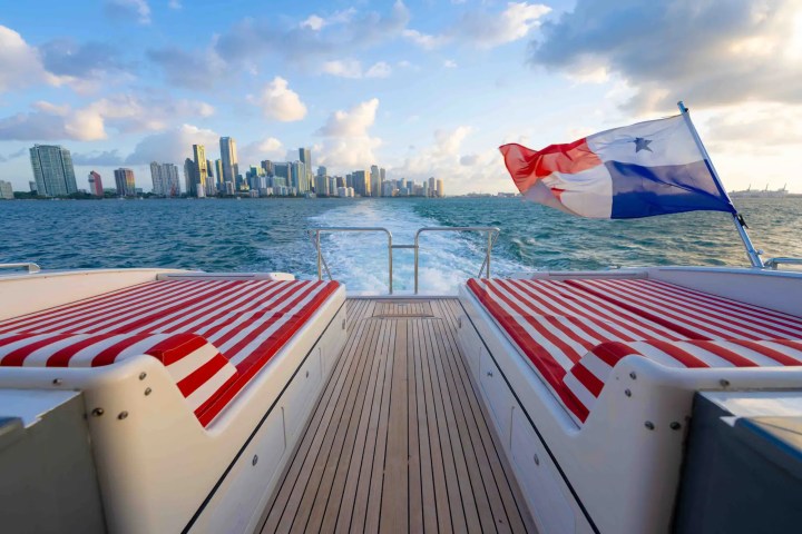 View from boat with red-striped seats and flag, city skyline in background on a sunny day.
