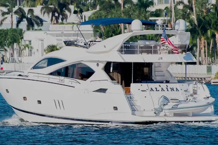 White yacht with blue canopy on water, American flag at the back, palm trees in background.