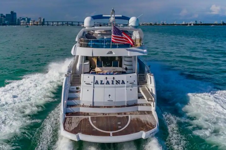 A large yacht with an American flag cruising in open water near a city skyline.