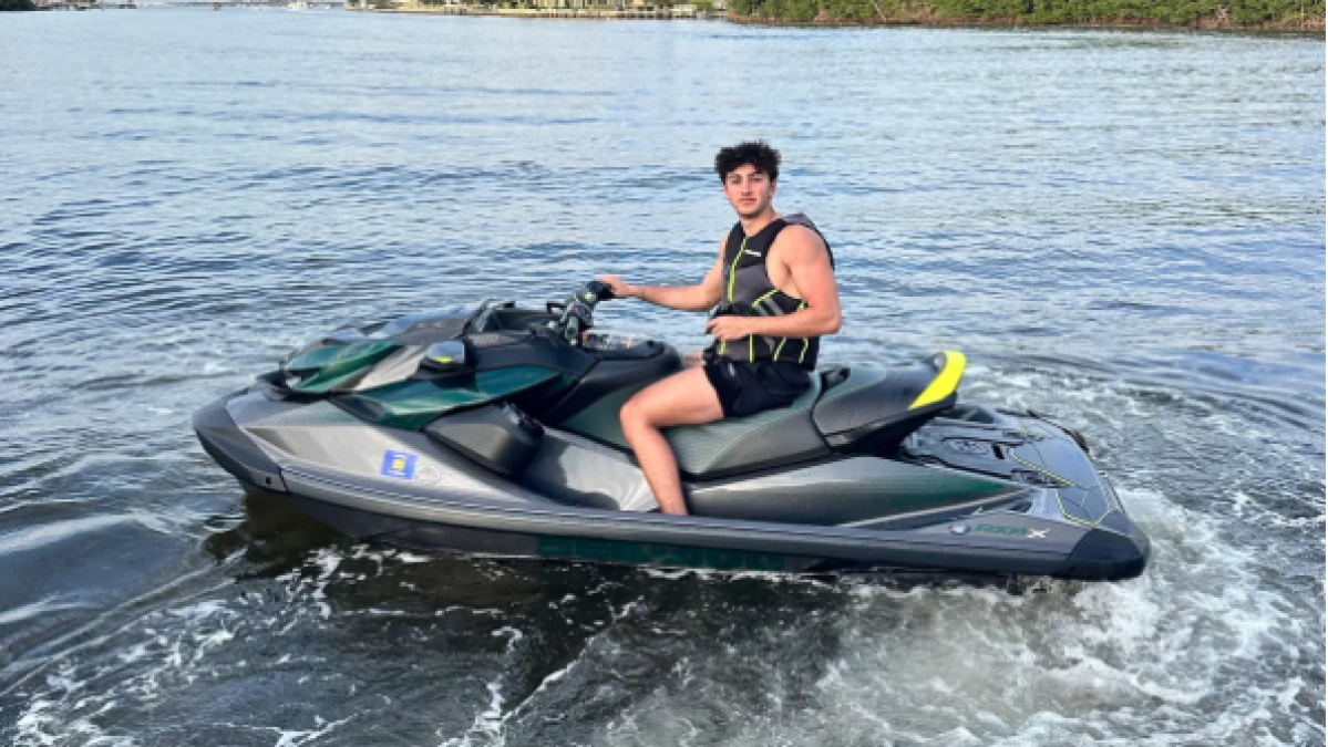 Person riding a jet ski on a calm body of water with trees and buildings in the background.