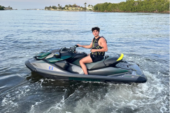 Person riding a jet ski on a calm body of water with trees and buildings in the background.