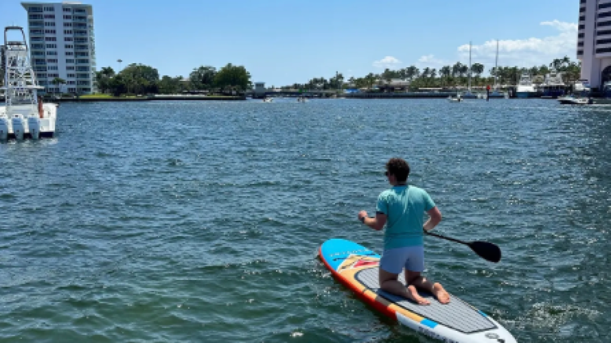 Person kneeling on a paddleboard in a calm waterway near tall buildings.