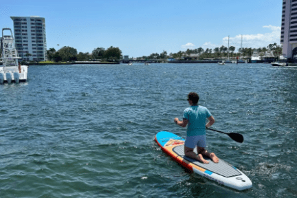 Person kneeling on a paddleboard in a calm waterway near tall buildings.