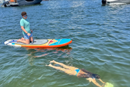 Person kneeling on paddleboard next to swimmer in ocean, with boats and city skyline in the background.