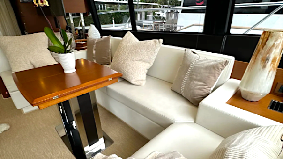 Luxury yacht interior with white seating, pillows, wooden table, and large windows showing outdoor view.