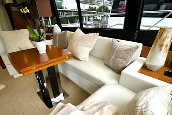 Luxury yacht interior with white seating, pillows, wooden table, and large windows showing outdoor view.