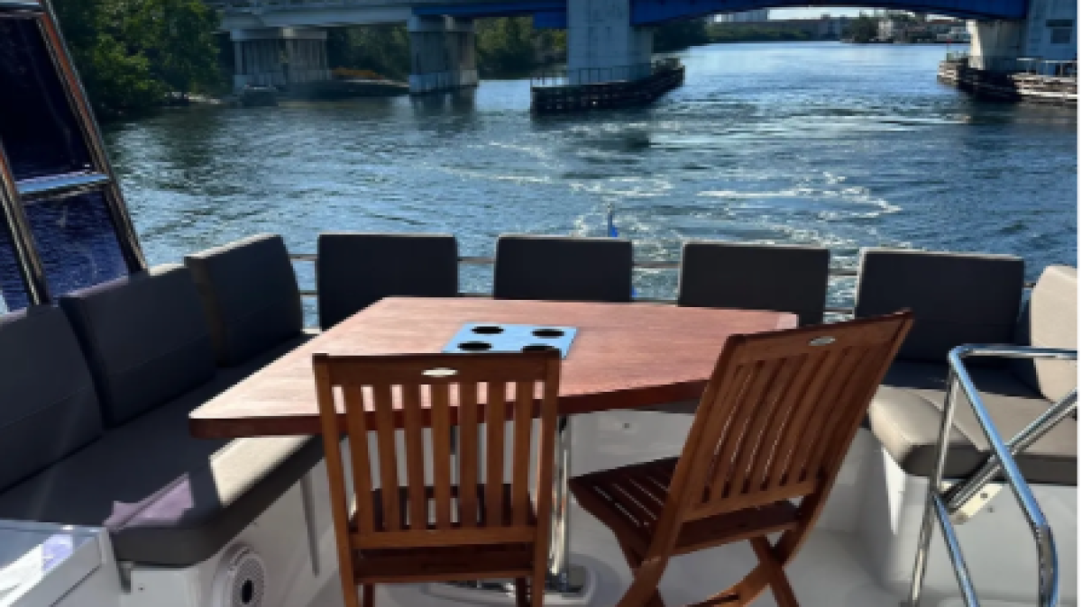 Boat deck with chairs and table, overlooking waterway and blue bridge under a sunny sky.