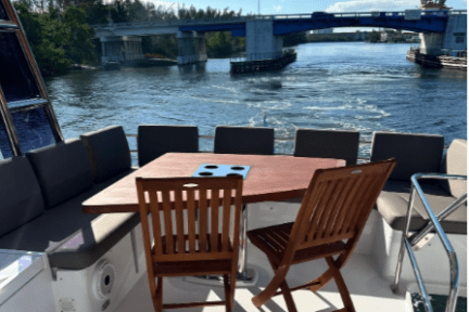 Boat deck with chairs and table, overlooking waterway and blue bridge under a sunny sky.
