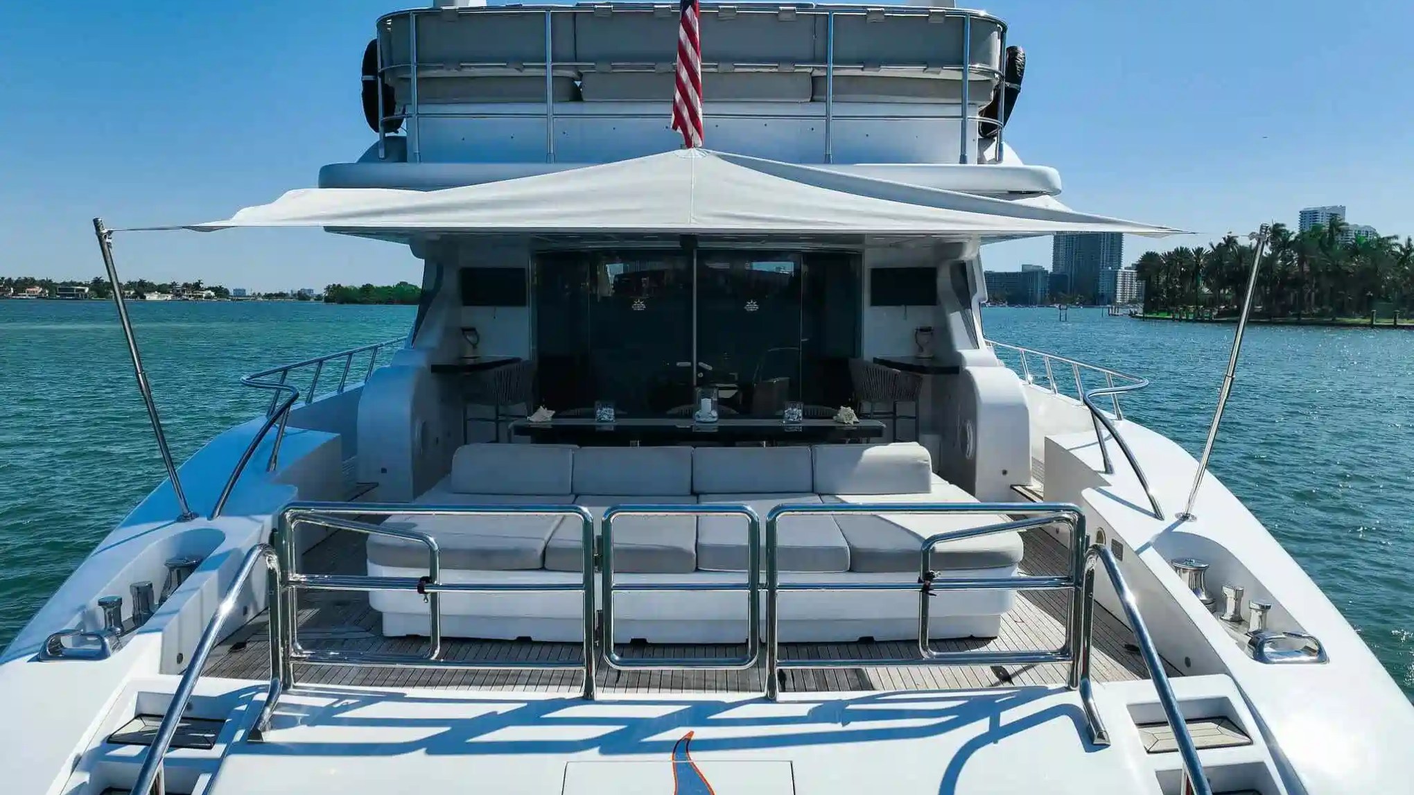 Rear deck of a luxury yacht with seating, canopy, and American flag on a sunny day.