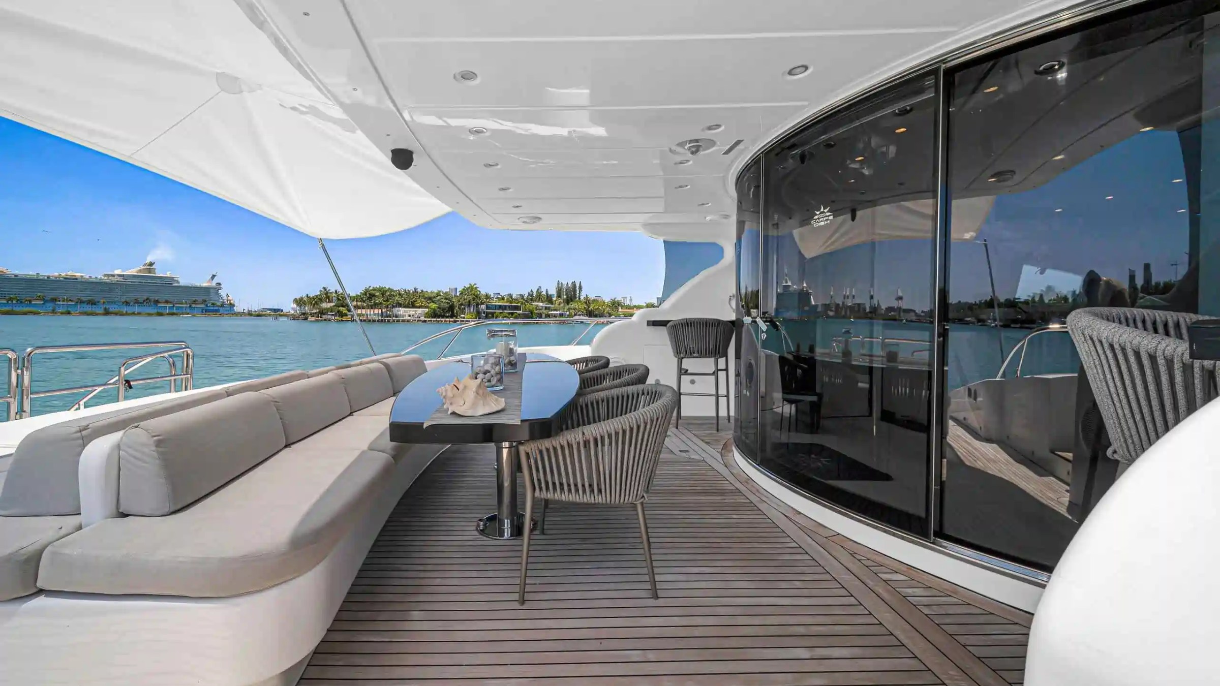 Luxury yacht deck with sofas, table, and ocean view; cruise ship and islands in background.