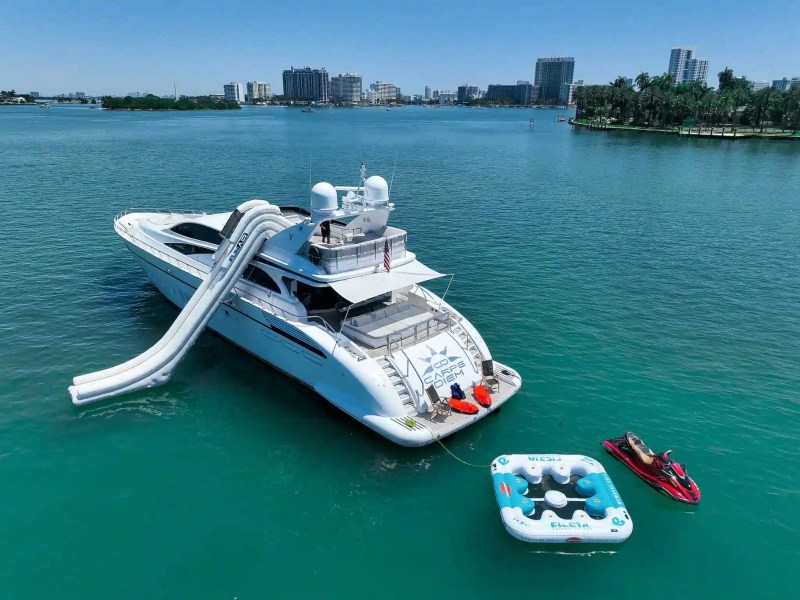 Yacht on water with inflatable slide, floating lounges, and jet ski nearby.