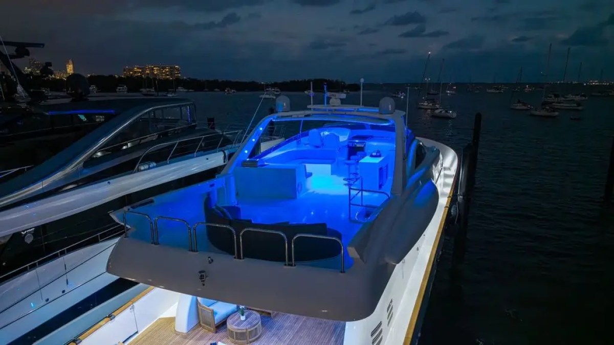 Luxurious yacht illuminated with blue lights at night, docked near a cityscape.