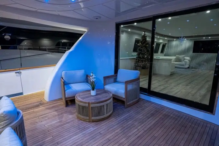 Yacht deck with wicker chairs, round table, indoor view of sofa, and Christmas tree through glass doors.