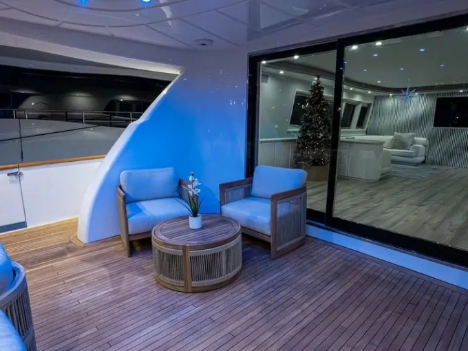 Yacht deck with wicker chairs, round table, indoor view of sofa, and Christmas tree through glass doors.
