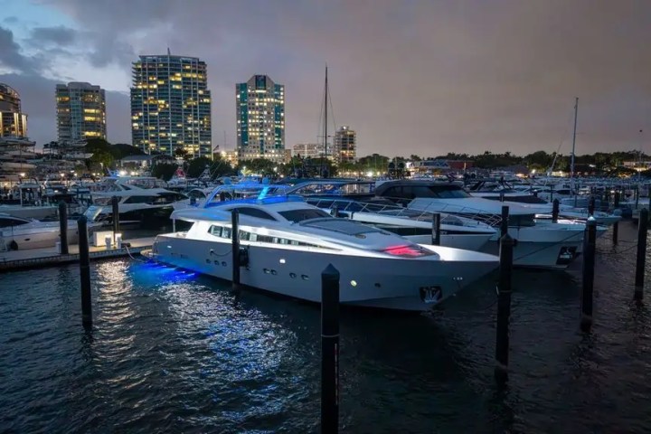 Yachts docked at a marina at dusk with illuminated city skyline in the background.