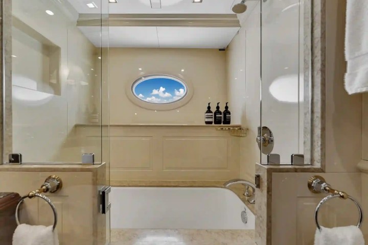 Luxurious bathroom with a bathtub, glass shower doors, and an oval window showing blue sky with clouds.