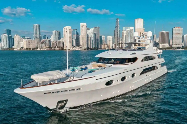 Luxury yacht sailing with a city skyline in the background under a clear blue sky.