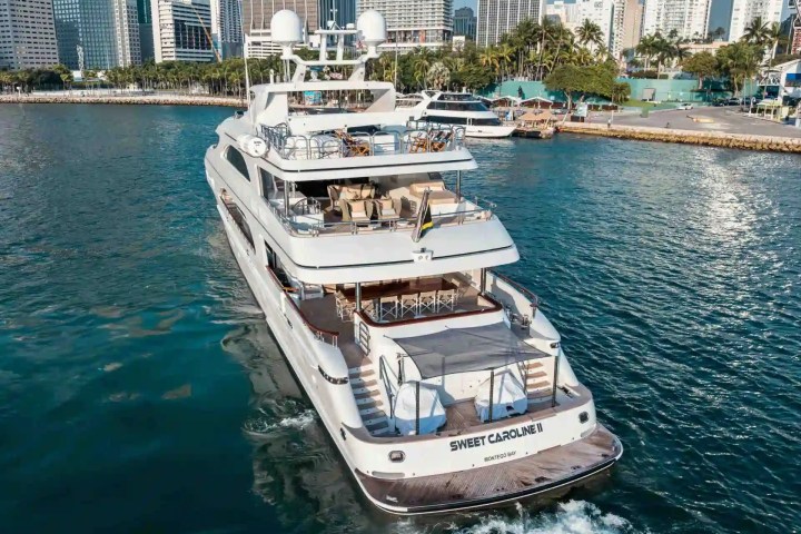 A luxury yacht named 'Sweet Caroline II' sails in a city harbor with skyscrapers in the background.