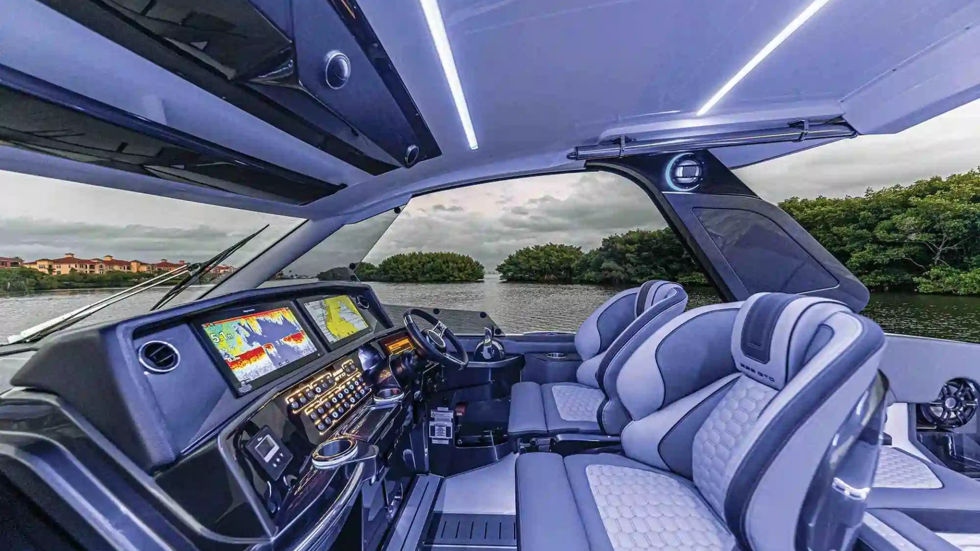 Luxury boat interior with high-tech dashboard and plush seating, set on a river with trees and buildings in view.