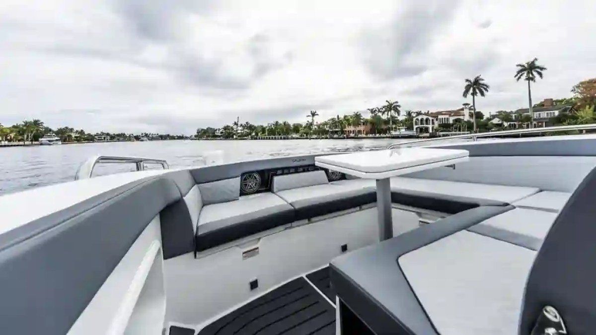 Luxury boat interior with gray seating and a table, overlooking a waterfront with palm trees and houses.