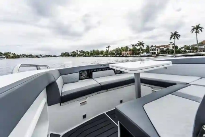 Luxury boat interior with gray seating and a table, overlooking a waterfront with palm trees and houses.