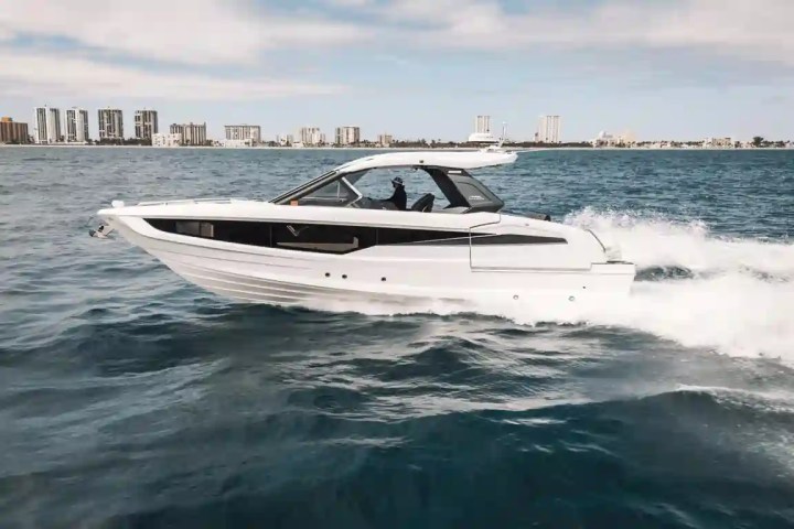 White speedboat cruising on blue ocean near city skyline under cloudy sky.