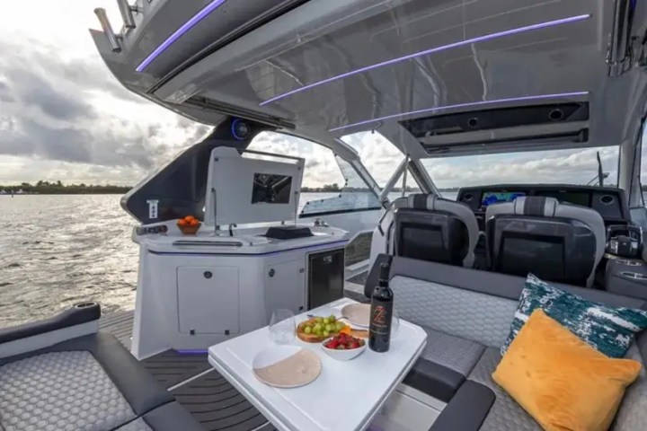 Luxury yacht interior with table, wine, fruit, and cushions, overlooking water.