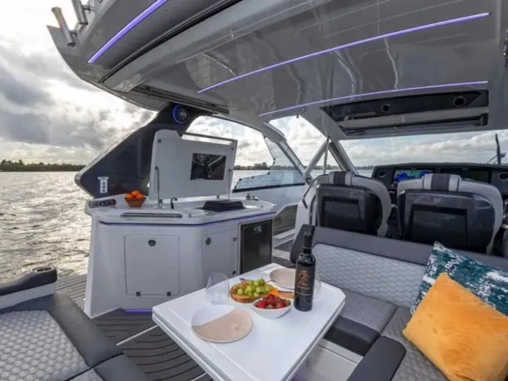 Luxury yacht interior with table, wine, fruit, and cushions, overlooking water.