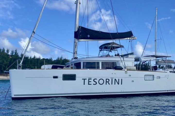White catamaran named Tesorini moored in blue water near a forested shoreline.