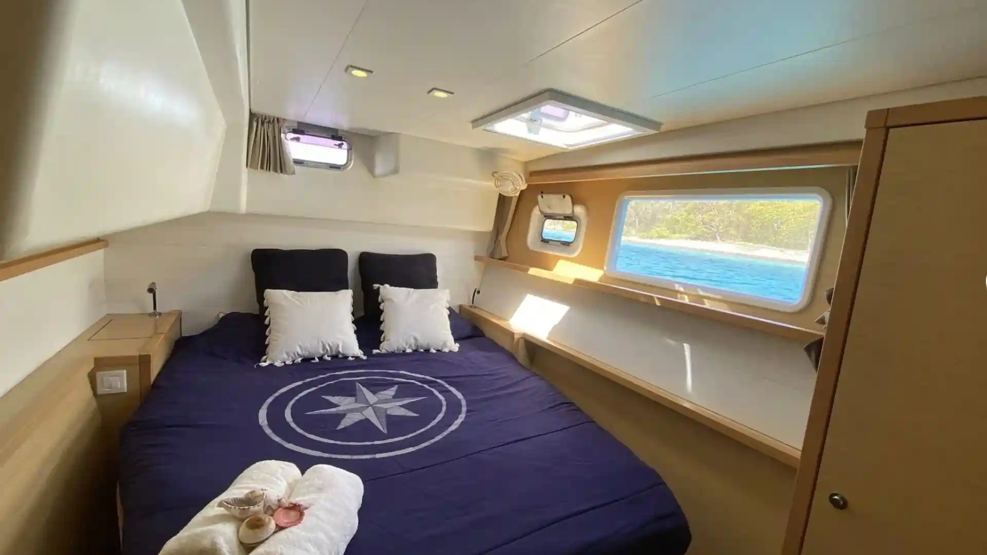 Cozy yacht cabin with a bed, navy bedding, and side windows overlooking the sea.