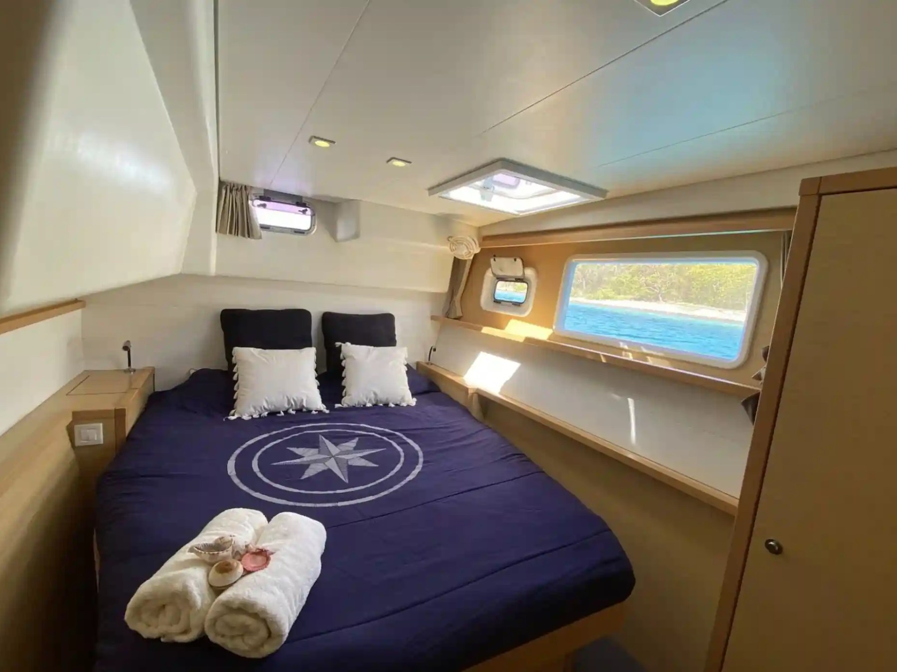 Cozy yacht cabin with a bed, navy bedding, and side windows overlooking the sea.