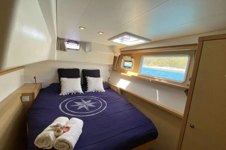 Cozy yacht cabin with a bed, navy bedding, and side windows overlooking the sea.