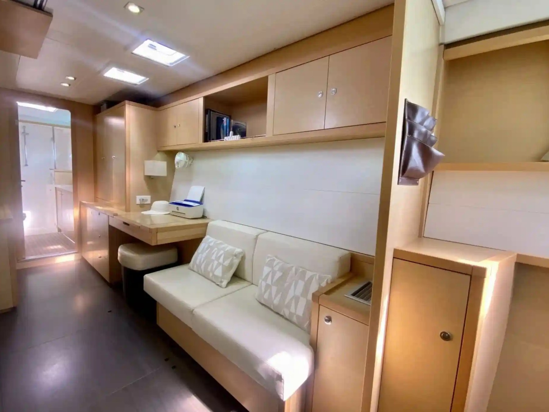 Modern yacht interior with beige sofa, desk, shelves, and doorway to a bright bathroom.