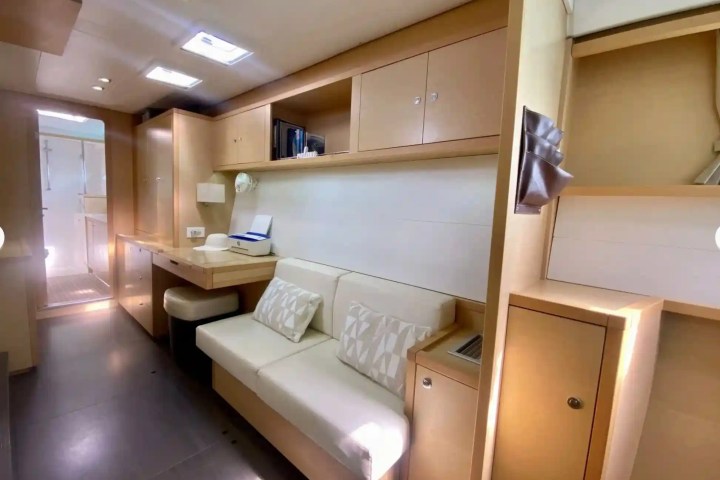 Modern yacht interior with beige sofa, desk, shelves, and doorway to a bright bathroom.