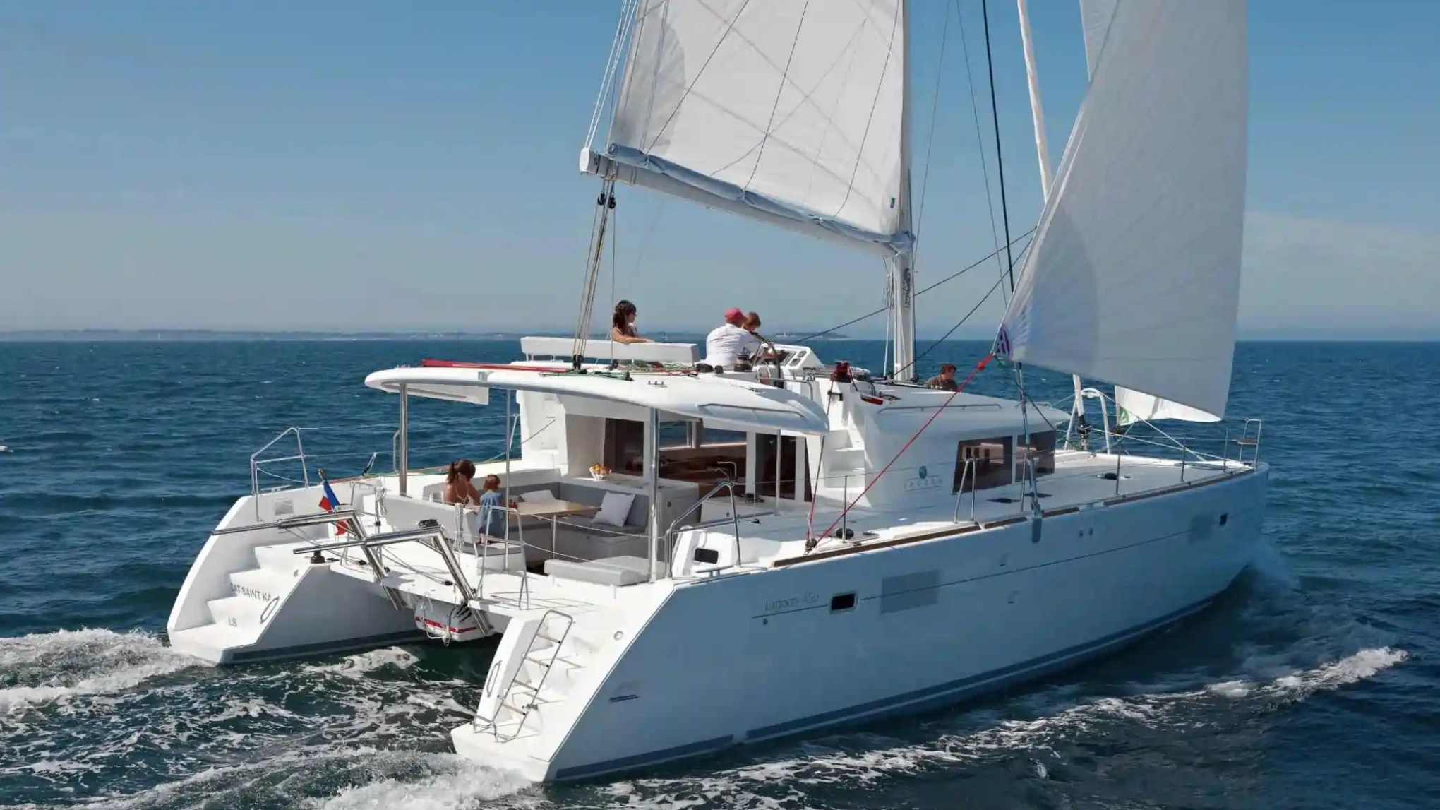 White catamaran sailing on the ocean with people on deck under clear blue skies.
