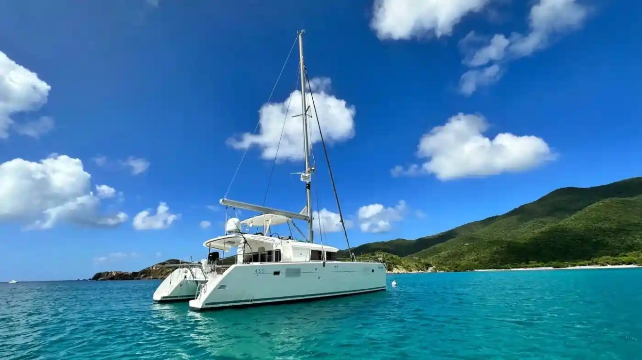 Sailboat on turquoise sea with cloudy sky and green hills in background.