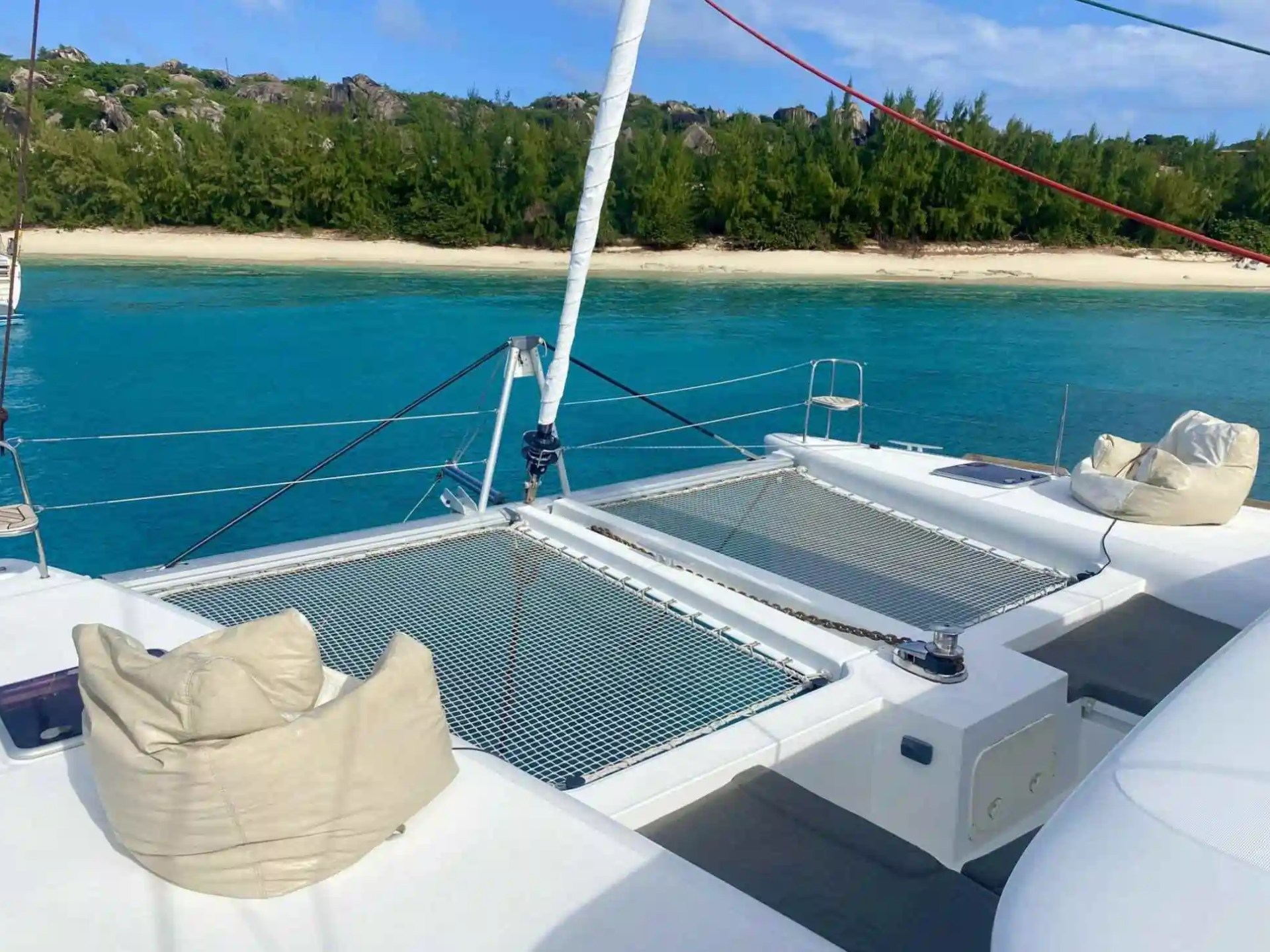View from a catamaran with netting, cushions, and turquoise water near a sandy beach and green trees.