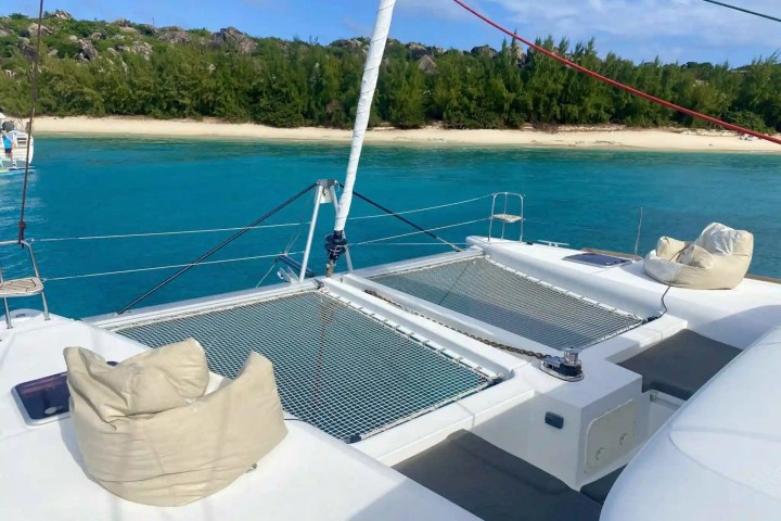 View from a catamaran with netting, cushions, and turquoise water near a sandy beach and green trees.